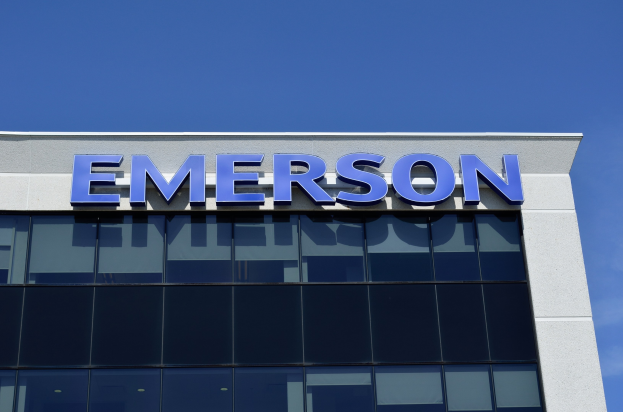Das Emerson-Logo an der Seite eines Gebäudes mit klarem blauem Himmel im Hintergrund, Text lautet "Emerson investiert 1,5 Milliarden US-Dollar in den USA".