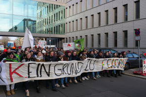 Eine Gruppe von Menschen auf der Straße mit einem Banner, auf dem "Kein Grind Lo Feuer" steht, im Protest, mit Gebäuden, Bäumen, Pfählen, Schildern, Fahrzeugen und einem klaren blauen Himmel im Hintergrund.