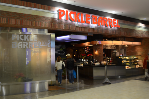 Ein Pickle-Bar-Restaurant in einem Einkaufszentrum mit Menschen, einem Schild, einer Auslage, Pflanzen, Metallstangen und Deckenleuchten.