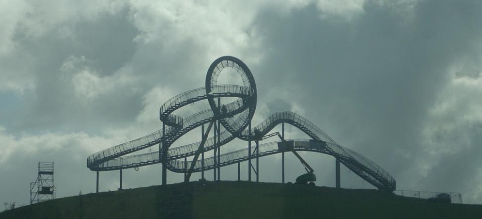 Eine Achterbahn auf dem Höhepunkt eines Hügels unter einem bewölkten Himmel, umgeben von Stützpfählen und -strukturen.