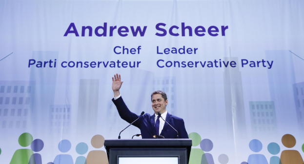 Konservative-Parteichef Andrew Scheer vor einem Podium mit zwei Mikrofonen und einem Bildschirm, vor einem Banner mit Text und Gebäudeabbildungen.