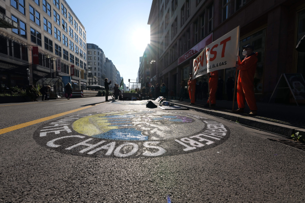 Eine Gruppe von Menschen mit Schildern und Plakaten auf einer Straße mit Gebäuden und Pflanzen, mit "Chaos" auf der Straße gemalt, während einer Protestaktion, die likely mit Occupy Wall Street in Verbindung steht.