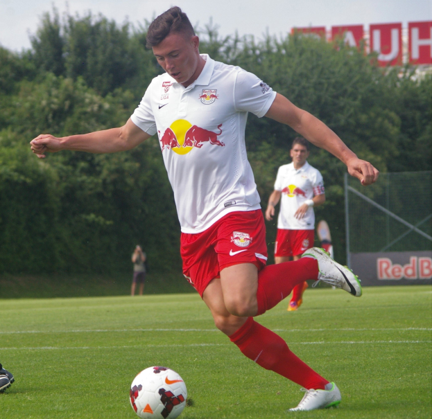 Ein professioneller Fußballspieler, der einen Ball auf einem grünen Feld mit Bäumen und einem klaren blauen Himmel im Hintergrund kickt, mit einer Tafel mit der Aufschrift "RB Leipzig" im Bild.