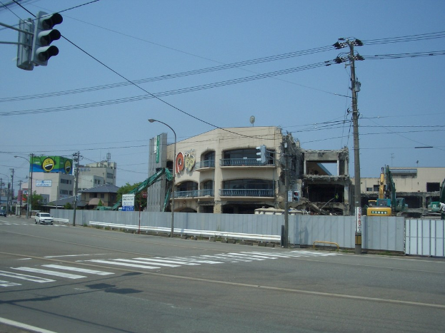 Eine Stadtstraße mit Fahrzeugen, Strommasten, Laternen, einer Ampel auf der linken Seite, Gebäuden, Bäumen und dem Himmel im Hintergrund sowie einem abbruchreifen Gebäude im Vordergrund.