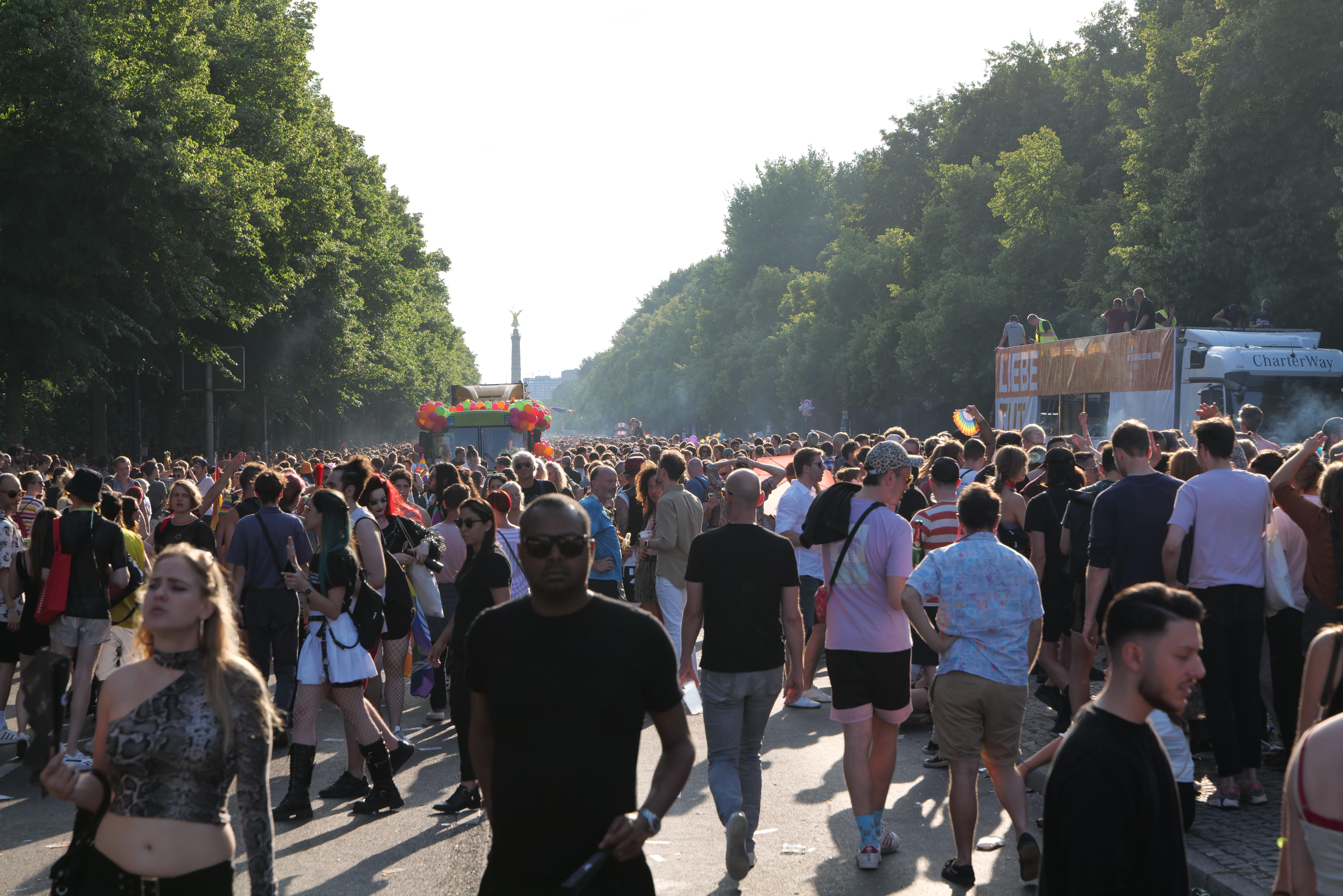 Eine große Menge von Menschen, die eine von Bäumen gesäumte Straße entlanggehen, mit einem Turm im Hintergrund und Fahrzeugen mit Teilnehmern auf der rechten Seite, wahrscheinlich beim Christopher Street Day in Berlin unter einem klaren blauen Himmel.
