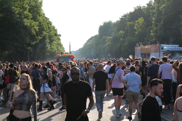 Eine große Menge von Menschen, die eine von Bäumen gesäumte Straße entlanggehen, mit einem Turm im Hintergrund und Fahrzeugen mit Teilnehmern auf der rechten Seite, wahrscheinlich beim Christopher Street Day in Berlin unter einem klaren blauen Himmel.