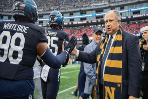 Ein Mann im Anzug und Krawatte gibt einem Fußballspieler die Hand auf einem Feld, umgeben von einer Gruppe von Menschen, mit einer Person, die eine Kamera auf der rechten Seite hält, und einem Stadion voller Menschen im Hintergrund, mit Schildern, die "Notre Dame Football Coach Mike McCarthy und die Fighting Irish" lesen.