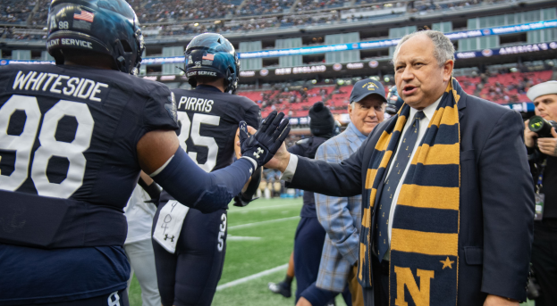 Ein Mann im Anzug und Krawatte gibt einem Fußballspieler die Hand auf einem Feld, umgeben von einer Gruppe von Menschen, mit einer Person, die eine Kamera auf der rechten Seite hält, und einem Stadion voller Menschen im Hintergrund, mit Schildern, die "Notre Dame Football Coach Mike McCarthy und die Fighting Irish" lesen.