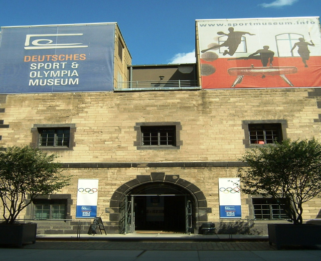 Außenansicht des Deutschen Sport & Olympia Museums in Berlin mit einem Gebäude mit Fenstern und einer Tür, umgeben von Bäumen und Bannern unter einem bewölkten Himmel.