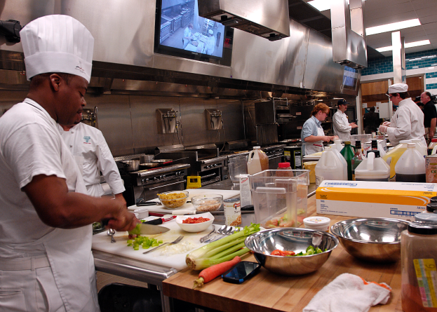 Eine Gruppe von Köchen in einer Küche, die auf einem Schneidbrett Essen zubereiten, wobei ein Koch im Vordergrund in einer weißen Schürze und einer Mütze steht und ein Messer hält und von verschiedenen Utensilien, Schüsseln, Gemüse, einem Handy, Flaschen und anderen Gegenständen umgeben ist, mit einem Fernseher und Deckenlampen im Hintergrund.