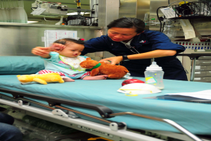 Eine Krankenschwester hält ein Baby in einem Krankenhauszimmer, mit Spielzeugen, einer Flasche und anderen Gegenständen auf dem Bett und medizinischen Geräten und einem Vorhang im Hintergrund.