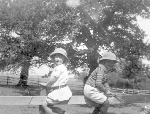 Zwei junge Jungs in Mützen mit Baseballschlägern in einem grasbewachsenen Park mit Bäumen und einem Zaun im Hintergrund, in Schwarz-Weiß.
