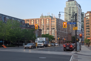 Eine belebte Stadtstraße mit verschiedenen Fahrzeugen wie einem Lastwagen, einem Auto und einem SUV, umgeben von Laternen, Ampeln, Texttafeln, Bäumen, Gebäuden mit Fenstern und einem Himmel im Hintergrund.