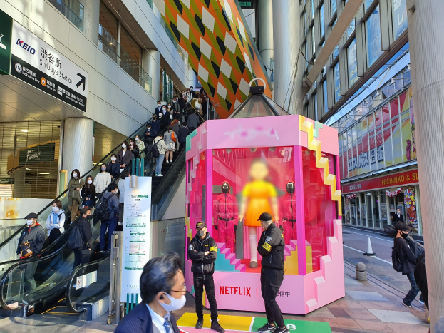 Eine Gruppe von Menschen steht vor einem Gebäude mit Glaswänden, Säulen und Texttafeln, einige tragen Masken, während eine Rolltreppe mit Menschen daneben aufsteigt; eine pinkfarbene Box in der Mitte zeigt Schaufensterpuppen und trägt 'Netflix' neue Netflix-Bühne in Tokio'.