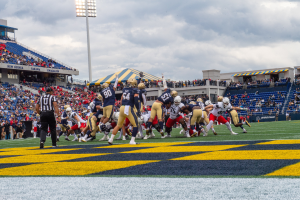 Ein Fußballspiel im Gange mit Spielern in Sportbekleidung und Helmen im Vordergrund, Zuschauern, Geländern, Beschriftungen, Laternenpfählen und einem bewölkten Himmel im Hintergrund und Text am unteren Bildrand.