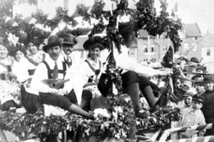 Eine Gruppe von Menschen in traditioneller bayrischer Tracht, einige mit Musikinstrumenten, fährt auf einemlaubgeschmückten Festwagen bei einem Umzug, mit Gebäuden und einem klaren Himmel im Hintergrund, dargestellt in Schwarz-Weiß.