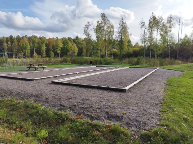 Eine grüne Wiese mit einem Picknicktisch in der Mitte, umgeben von Bäumen und einem klaren blauen Himmel, mit Spielplatzgeräten wie Stangen und einer Bank auf der linken Seite.