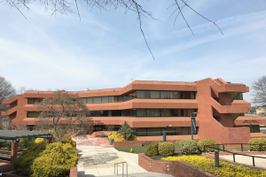 Das neue akademische Zentrum der Universität von Pennsylvania, ein großes Backsteingebäude mit einem Weg davor, umgeben von Bäumen, Pflanzen, Blumen, Gras, Geländern, Pfählen, Brettern und einem Schuppen, mit Fahrzeugen auf der Straße und einem bewölkten Himmel im Hintergrund.