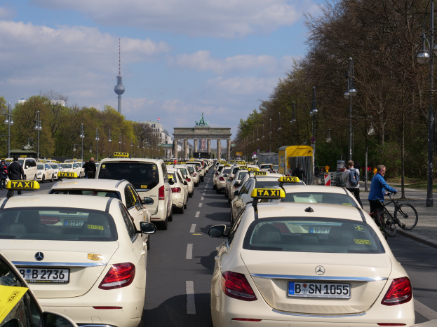 Eine lange Reihe von Taxis steht an der Seite einer belebten Straße in Berlin, Deutschland, mit Fahrzeugen, Radfahrern und Fußgängern, flankiert von Laternenmasten, Bäumen und Gebäuden, einschließlich eines Bogens und eines Turms.