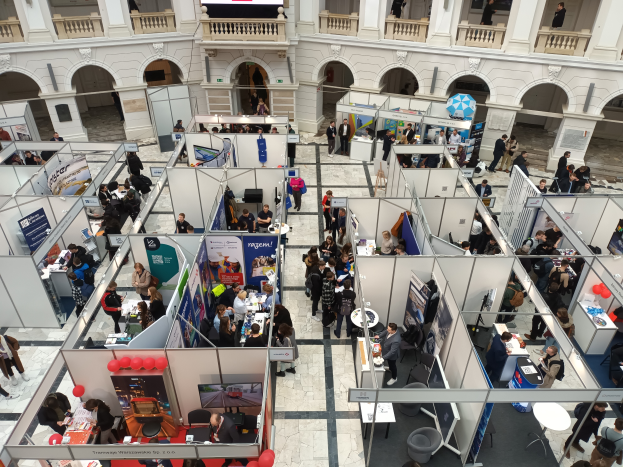 Eine große Gruppe von Menschen versammelte sich in einem Raum, der mit Ständen, Bannern, Tischen, Stühlen und Ballons gefüllt war, mit einem Gebäude im Hintergrund, das Bögen, Säulen, Geländer und einen Bildschirm aufwies.