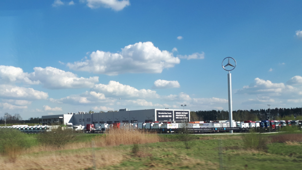Luftaufnahme eines Mercedes-Benz-Werks in Deutschland mit Fahrzeugen auf der Straße, Grünflächen, Industriegebäuden und einem logo tragenden Pfahl unter einem bewölkten Himmel.