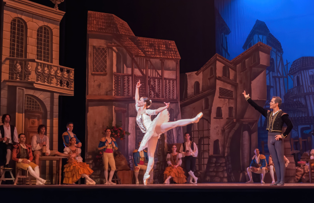 Zwei Tänzer performen auf einer Bühne im Royal Opera House in London, mit einem Publikum im Vordergrund, das auf Stühlen sitzt und steht, und einer projizierten Stadtkulisse im Hintergrund.
