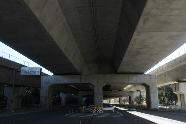 Autobahn unter einer Brücke mit Absperrkegeln, Barrieren und Texttafeln am Straßenrand, Geländern an den Seiten, Bäumen und einem klaren blauen Himmel im Hintergrund.