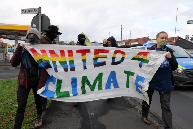 Eine Gruppe maskierter Personen mit einer 'United Climate'-Schleife vor einer Tankstelle, Autos auf der Straße, Gras, Schilder, Gebäude, Bäume und ein bewölkter Himmel im Hintergrund.