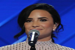 Demi Lovato steht selbstbewusst vor einem Mikrofon bei den Grammys, trägt ein schwarzes Kleid und eine stylische Bob-Frisur, vor einem blauen Hintergrund.