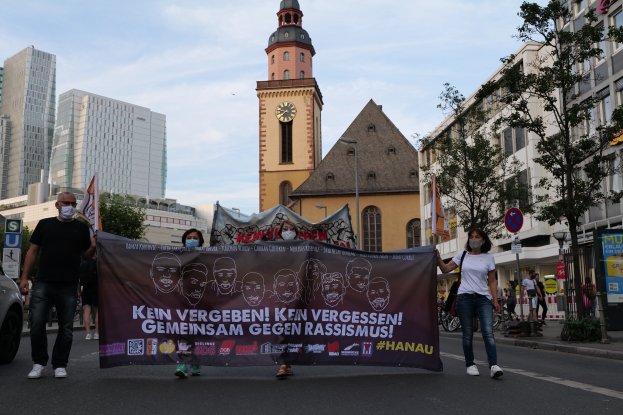 Eine Gruppe maskierter Menschen marschiert auf einer Straße und hölt ein Banner mit Text und Bildern. Auf der linken Seite des Bildes steht ein Auto auf der Straße. Im Hintergrund sind Gebäude, Bäume, Schilder, Masten und ein Uhrturm zu sehen.