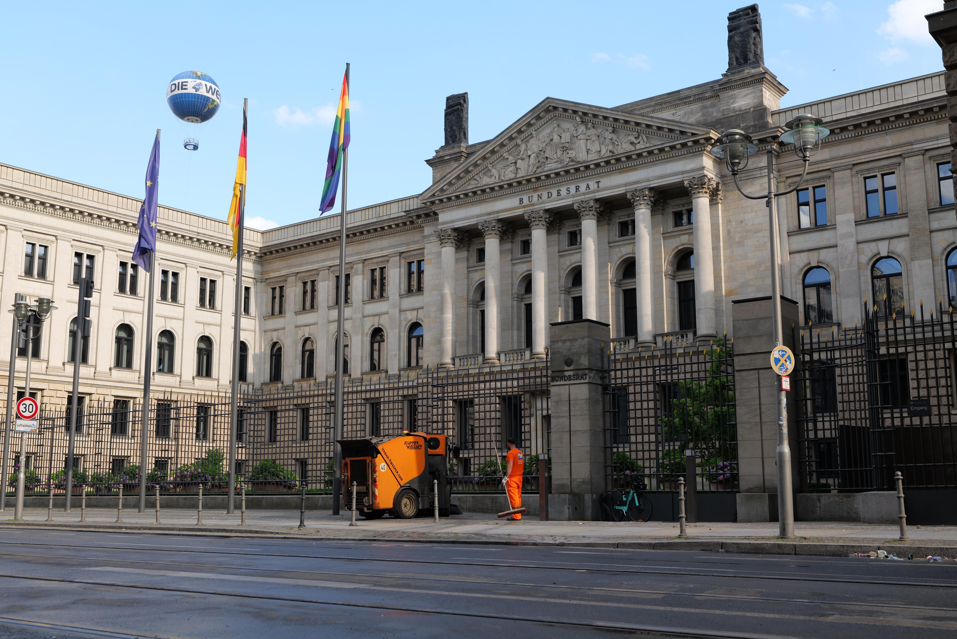 Großes modernes Gebäude mit vielen Fenstern, als Bundestag in Berlin identifiziert, umgeben von Straßeninfrastruktur, Fußgängern, Fahrzeugen, Flaggen und Bäumen unter einem bewölkten Himmel.