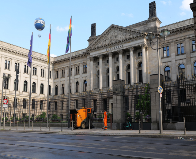 Großes modernes Gebäude mit vielen Fenstern, als Bundestag in Berlin identifiziert, umgeben von Straßeninfrastruktur, Fußgängern, Fahrzeugen, Flaggen und Bäumen unter einem bewölkten Himmel.