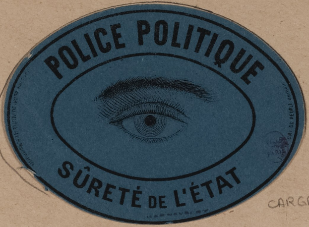 Brauner Karton mit blauem Aufkleber mit der Aufschrift 'Polizei Politique Sûreté de l'État' und einem Auge-Symbol.
