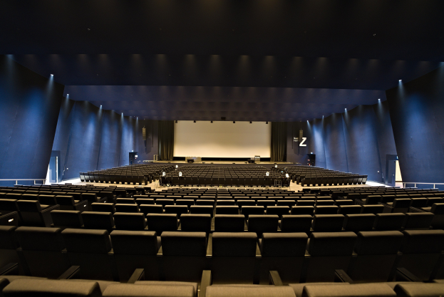 Ein großer Zuschauerraum mit Reihen leerer Stühle, ein Projektionsschirm und Bühnenobjekte, beleuchtet von Wandlampen.