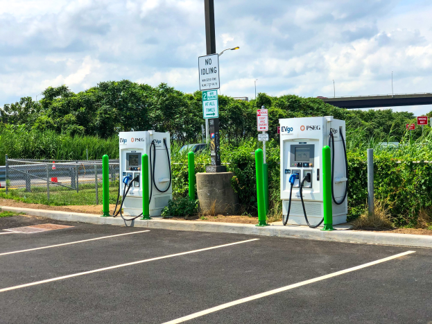 Elektroauto-Ladestation auf einem Parkplatz mit umgeben-der Infrastruktur, Vegetation und einer Brücke im bewölkten Himmelhintergrund.