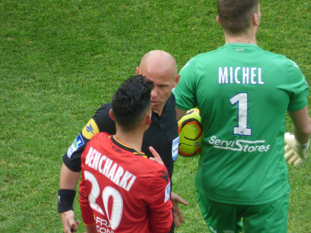 Schiedsrichter spricht mit zwei Fußballspielern auf einem Rasenfeld, ein Spieler hält einen Ball.