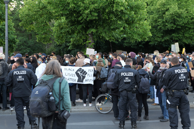 Eine große Gruppe von Menschen steht auf der Straße und hält Schilder und trägt Mützen und Taschen, mit einem Fahrrad im Vordergrund und Bäumen und einem Pfahl im Hintergrund während einer Black-Lives-Matter-Demonstration in Berlin.