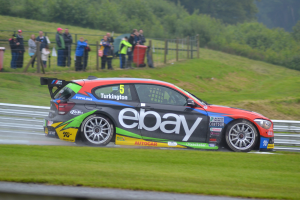Ein Auto, das Rauch beim Fahren auf einer Rennstrecke ausstöüst, umgeben von Gras, einem Zaun, Mülltonnen, Menschen, Bäumen und einem klaren blauen Himmel.