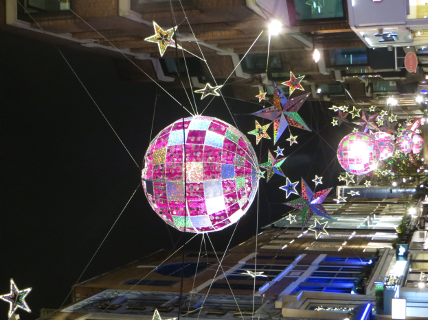 Eine Stadtstra├če bei Nacht mit zahlreichen Lichtern und Deko, mit einem gro├čen Diskokugel, die von einem Geb├Ąude h├Ąngt und eine festliche Atmosph├Ąre schafft.