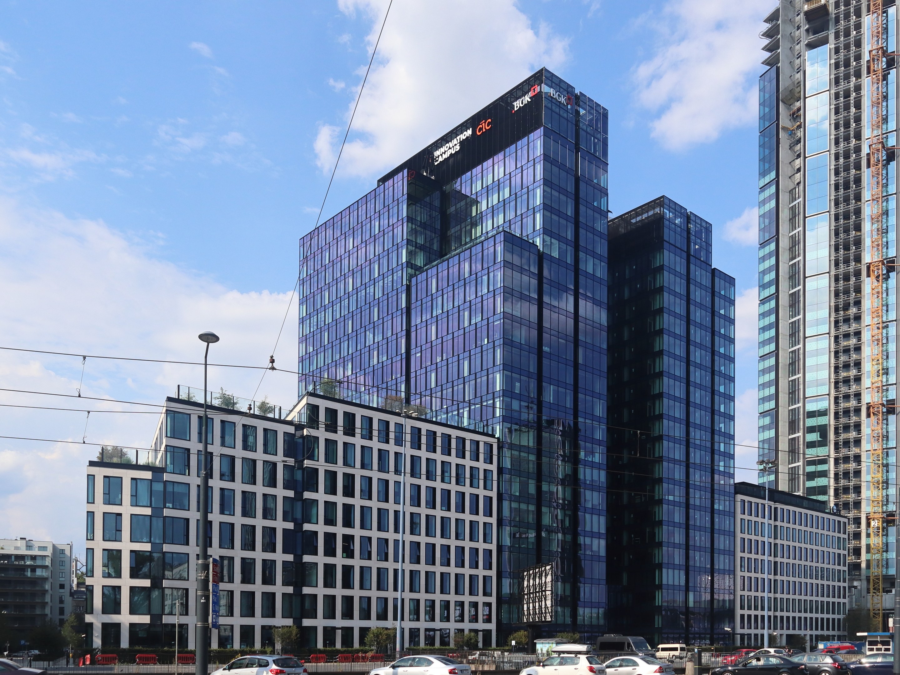 Zentrale der Deutschen Bank in Frankfurt, Deutschland, ein großes mehrfenstriges Gebäude umgeben von städtischer Infrastruktur einschließlich Straßenlaternen, Elektro Polen, Elektrokabel, Kraftfahrzeugen auf der Straße, Büschen, Bäumen und einem wolkenverhangenen Himmel.