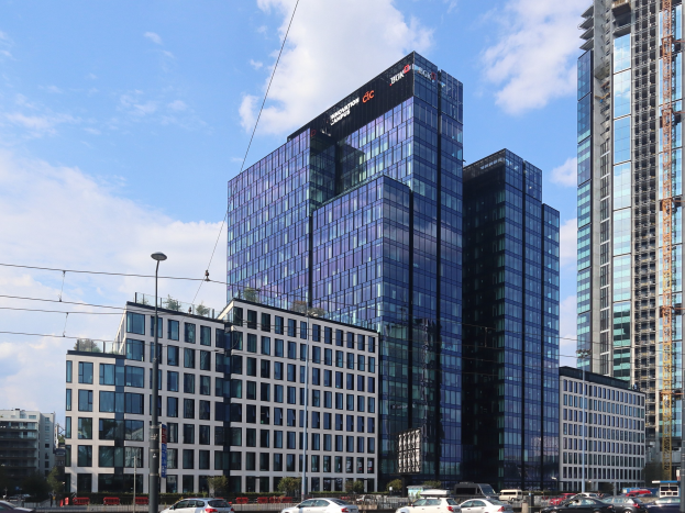 Zentrale der Deutschen Bank in Frankfurt, Deutschland, ein großes mehrfenstriges Gebäude umgeben von städtischer Infrastruktur einschließlich Straßenlaternen, Elektro Polen, Elektrokabel, Kraftfahrzeugen auf der Straße, Büschen, Bäumen und einem wolkenverhangenen Himmel.