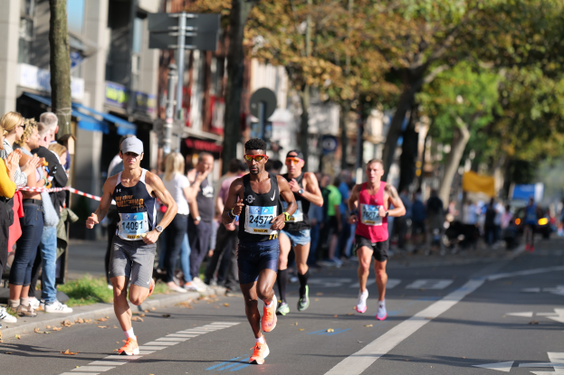 Eine Gruppe von Menschen, die bei einem Marathon auf einer Stadtstraße laufen, mit Zuschauern auf der linken Seite, Bäumen, Gebäuden und einem klaren blauen Himmel im Hintergrund. Alle Läufer tragen Turnschuhe und Hüte.