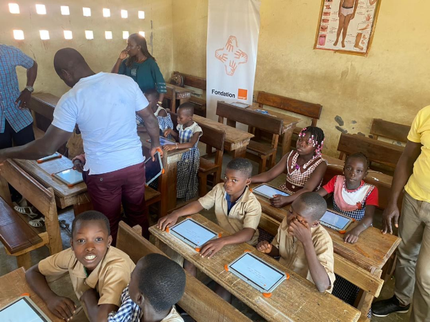 Kinder an Tischen sitzend, die Tablets verwenden, in einem Klassenzimmer mit Erwachsenen in der Nähe, einem Banner und einem Plakat an der Wand.