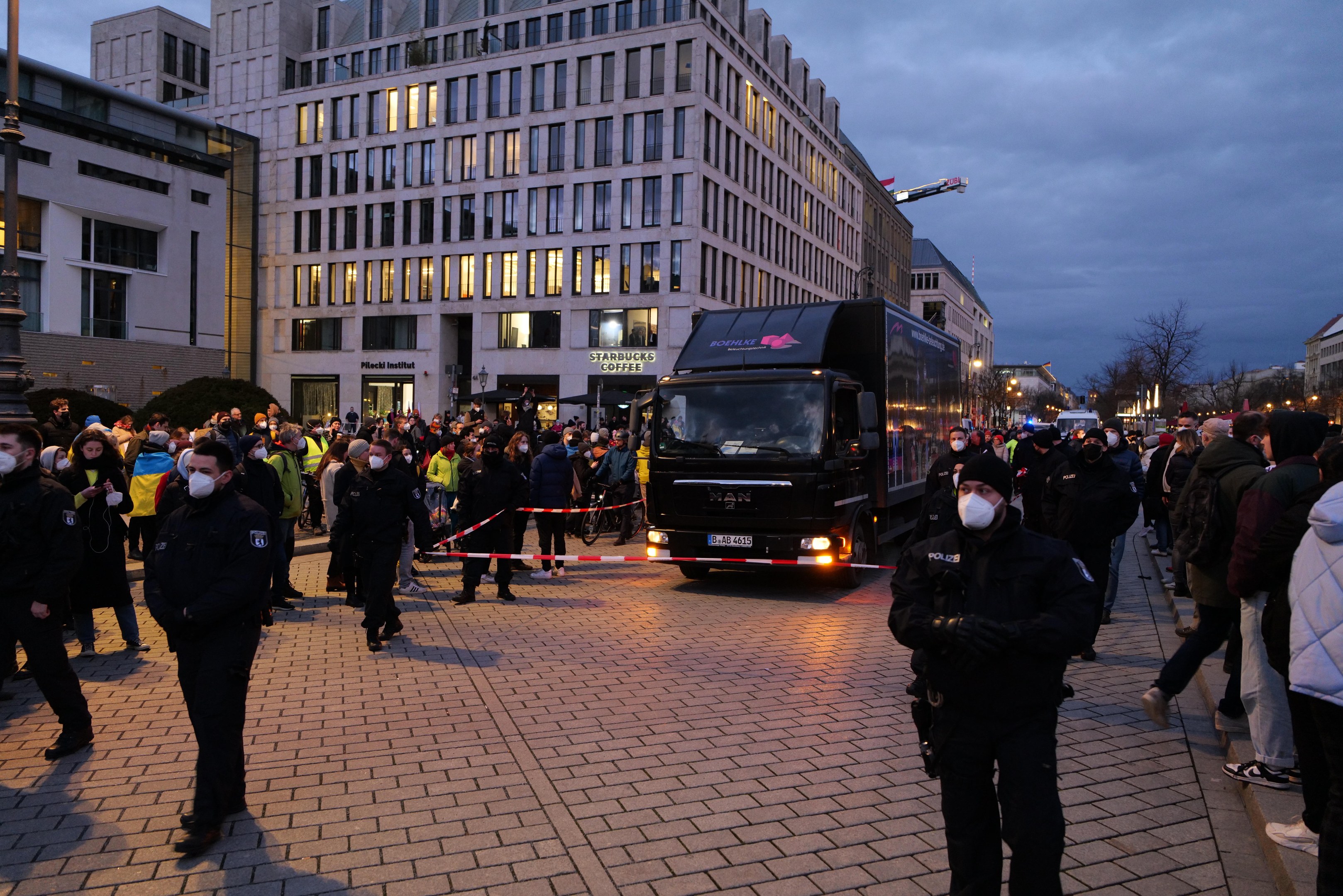 Gruppe von Menschen vor einem Lastwagen auf einer Straße mit Gebäuden, Bäumen und Laternen im Hintergrund, einige tragen Mützen und Masken, mit einem Band an einem Pfahl im Vordergrund.