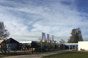Ein große Gebäude mit einer grünen Rasenfläche davor, umgeben von einem Metallzaun, einem Weg, Bäumen, Masten, Bannern, einem geparkten Fahrrad, näheren Häusern und einem bewölktem Himmel, das als neuer Spielplatz für Kinder an einer Schule dient.
