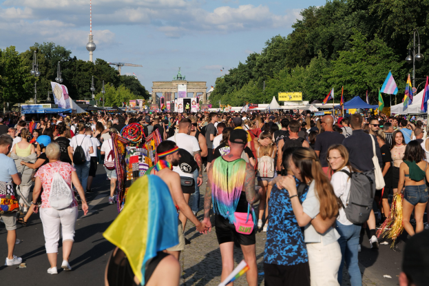 Große Menschenmenge auf der Straße in Berlin, Deutschland, viele tragen Taschen und halten Fahnen, mit Zelten an den Seiten, Bäumen, Laternenmasten, einem Bogen, einem Turm und einem bewölkten Himmel im Hintergrund.