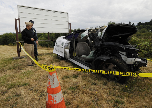 Ein Polizist in Uniform steht neben einem schwer beschädigten Auto am Straßenrand, mit eingedellter Motorhaube und zersplitterten Fenstern, neben einem Verkehrskegel und einer Banderole an einem Pfahl, mit Bäumen und einem bewölkten Himmel im Hintergrund.