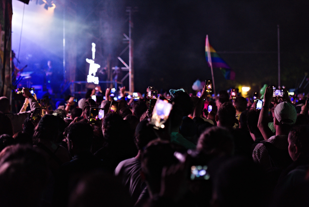 Menge an Menschen auf einem Konzert mit hochgehaltenen Handys, beleuchtet von Lichtern und umgeben von Pfählen und einer Flagge, während Musiker auf der Bühne spielen.
