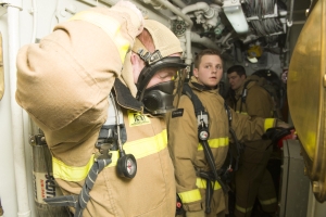 Feuerwehrleute in Gasmasken und Schutzausrüstung stehen in einem Raum mit Rohren und Gegenständen, mit einem Metallobjekt auf der rechten Seite.