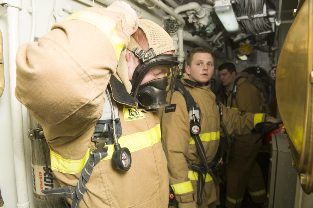Feuerwehrleute in Gasmasken und Schutzausrüstung stehen in einem Raum mit Rohren und Gegenständen, mit einem Metallobjekt auf der rechten Seite.
